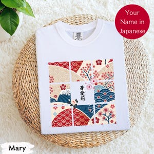 以下が含まれることがあります： 日本のデザインの白いTシャツ。グラフィックは、桜と波のある赤、ベージュ、青のパターンのグリッドを特徴としています。日本語のテキストは中央に配置され、赤い円には「あなたの名前（日本語）」と書かれています。