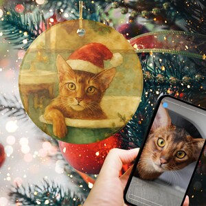 以下が含まれることがあります： サンタ帽をかぶった猫の水彩画が描かれた丸いクリスマスオーナメント。猫は茶色で、黄色い目をしています。オーナメントは金の紐から吊り下げられています。スマートフォンには同様の猫の画像が表示されています。