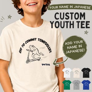 Puede incluir: Camiseta juvenil personalizada de color crema con un gráfico negro de una rana en patinete y el texto "OFF TO COMMIT TOMFOOLERY". Incluye los textos "YOUR NAME IN JAPANESE" y "ADD YOUR NAME IN JAPANESE!".