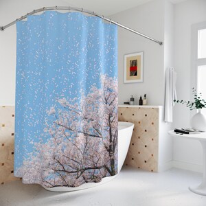 Op de afbeelding: Douchegordijn met een blauwe lucht en kersenbloesem ontwerp. Het gordijn hangt aan een gebogen stang in een badkamer. Het ontwerp toont een boom met roze en witte bloemen en vallende bloemblaadjes.