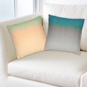 Peut inclure: Deux coussins carrés avec des motifs ondulés. L'un présente un dégradé de vert à pêche, l'autre de turquoise à gris. Les coussins sont posés sur un canapé en cuir blanc.