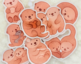 May include: A set of nine adorable otter stickers in various poses, including holding a heart, a fish, and cuddling. The stickers are painted in a soft, watercolor style with peach and brown tones.