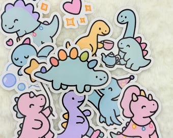 May include: A set of nine colorful dinosaur stickers with various designs, including a dinosaur holding a teacup, a dinosaur with a party hat, and a dinosaur with a star. The stickers are on a white fuzzy background.