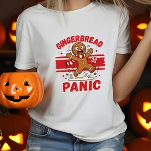 May include: A white short-sleeved t-shirt featuring a graphic of a panicked gingerbread character with a red and white scarf, surrounded by red text that reads GINGERBREAD PANIC, with a small peppermint candy and red horizontal stripes.