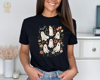 May include: Black t-shirt featuring a circular design of white cartoon ghosts with various expressions, surrounded by colorful flowers and foliage. A bat and crown are also included in the design. The shirt is a crew neck.