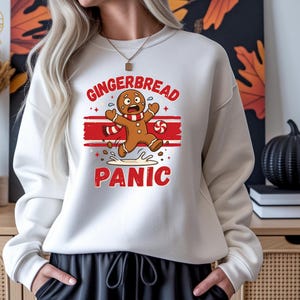 May include: A white crewneck sweatshirt featuring a graphic of a gingerbread person with a panicked expression, wearing a red and white striped scarf. The text GINGERBREAD PANIC is printed in red above and below the character with red stripes.
