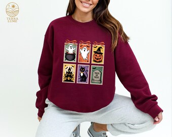 May include: Burgundy sweatshirt featuring six Halloween-themed postage stamp designs. The stamps depict a ghost in a cauldron, a ghost, a jack-o'-lantern, a haunted house, a black cat, and a tombstone with "Trick or Treat" text. The sweatshirt has a crew neck.