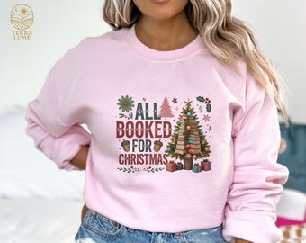 May include: A light pink sweatshirt with the text "ALL BOOKED FOR CHRISTMAS" in a square design. The design includes a Christmas tree made of books, gifts, and festive elements.