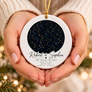 May include: A round white ceramic ornament featuring a circular dark blue night sky graphic with gold star constellations. The text reads Robert & Sophia, THE NIGHT WE MET, APRIL 9TH, 2021, AUSTIN, TEXAS, and 30.2672 N 97.7431 W with a gold hanging cord.