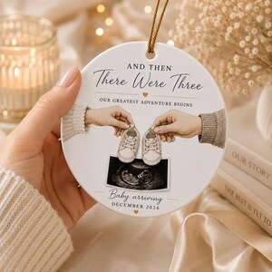 May include: A white ceramic disc ornament with gold string. It features the text "AND THEN There Were Three" and "OUR GREATEST ADVENTURE BEGINS." An ultrasound image and the text "Baby arriving DECEMBER 2026" are also present. Two hands hold baby shoes.