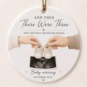 May include: Round white ceramic ornament with gold string, featuring the text "AND THEN There Were Three" and "OUR GREATEST ADVENTURE BEGINS". The ornament includes illustrations of baby shoes, an ultrasound image, and the text "Baby arriving OCTOBER 2025".