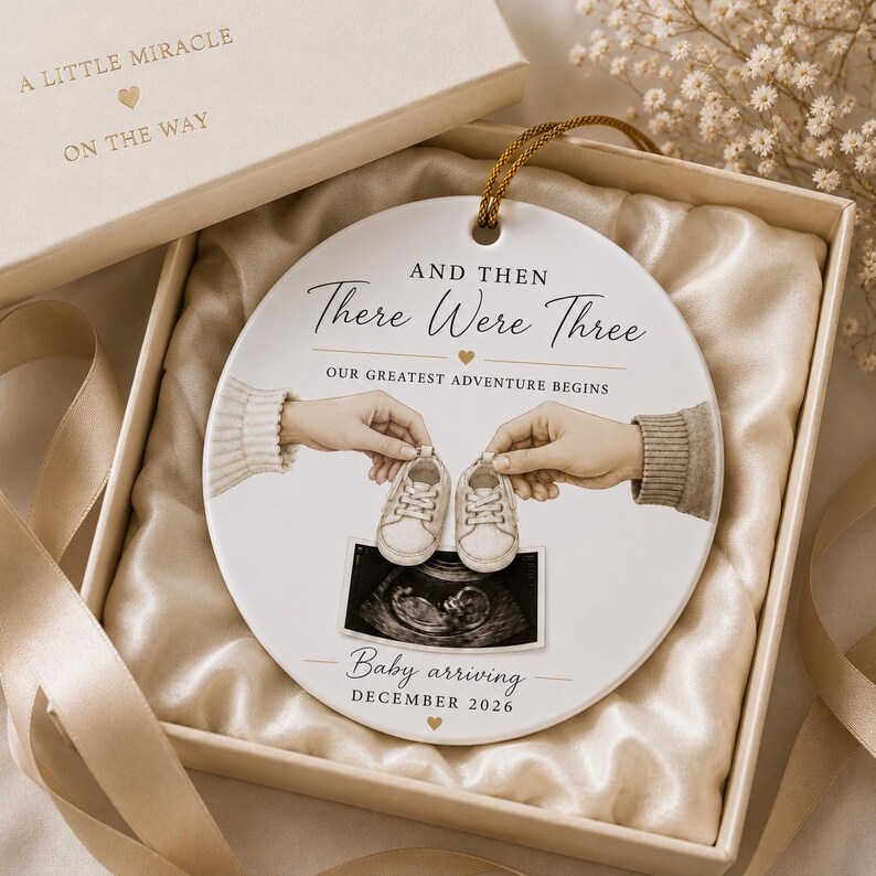 May include: A white ceramic disc ornament with gold detailing, inscribed with "And Then There Were Three" and "Baby arriving December 2026." The design includes baby shoes and an ultrasound image, presented in a gift box with a gold ribbon.