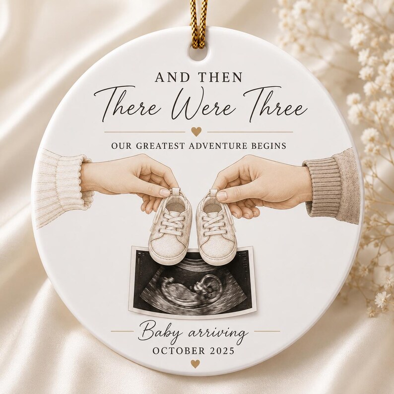 May include: A white ceramic ornament with a gold string features the text "And Then There Were Three" and "Our Greatest Adventure Begins." It depicts hands holding baby shoes above an ultrasound image. The text "Baby arriving October 2025" is also present.