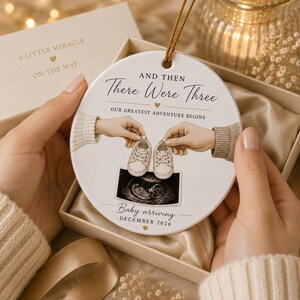 May include: A white ceramic ornament with gold text and a gold string. The ornament reads "And Then There Were Three" and "Baby arriving December 2026". The ornament features a sonogram image and two pairs of baby shoes.