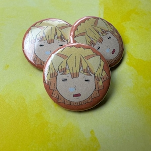 Può includere: Tre bottoni rotondi raffiguranti un personaggio dei cartoni animati con i capelli gialli e un'espressione triste. I bottoni hanno un bordo rosso-arancio e sono disposti su uno sfondo giallo brillante. Il volto del personaggio è dettagliato con accenti marroni e rossi.