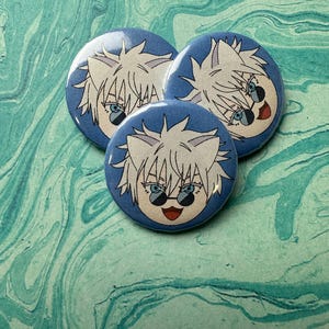 Può includere: Tre bottoni rotondi blu con un design di personaggio dei cartoni animati. Il personaggio ha i capelli bianchi, occhi blu e indossa occhiali da sole. I bottoni sono disposti su una superficie marmorizzata verde acqua e bianca.