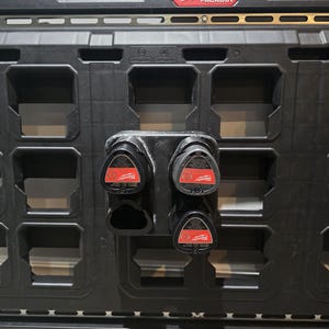 May include: Black Milwaukee Packout storage system with a red and white logo. The image shows a close-up of the storage system with a 3D-printed battery holder attached, holding four red and black batteries.