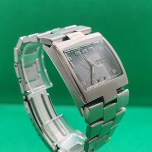 May include: A silver wristwatch with a rectangular face and a matching link strap. The watch face shows the date, with the text "MER 18" visible. The watch is set against a green backdrop.