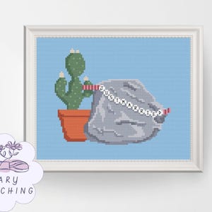 May include: Cross-stitch art featuring a green cactus in a terracotta pot next to a gray rock formation. A banner across the rock reads "JUST MARRIED" in black letters. The background is light blue, framed in white.