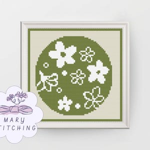 May include: A framed cross-stitch floral design. The artwork features white flowers on a green circular background. The frame is white. The logo "MARY STITCHING" is in the lower left corner.