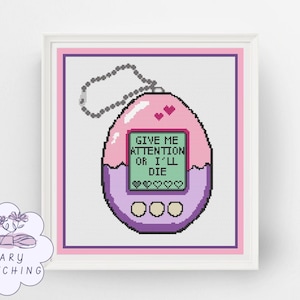 Puede incluir: Arte de punto de cruz de una mascota digital rosa y morada estilo Tamagotchi con el texto "GIVE ME ATTENTION OR I'LL DIE". El diseño incluye corazones y una cadena. La obra de arte está enmarcada.