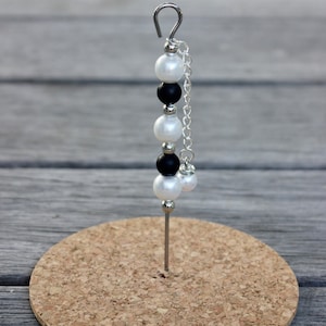 May include: A silver hair clip with a chain and black and white beads. The clip is on a cork surface.