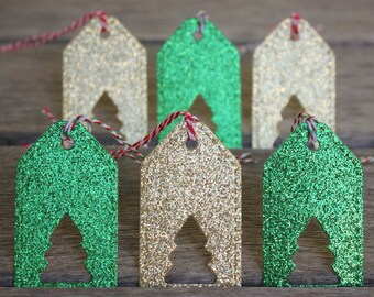 May include: Glittery gift tags in gold and green, each with a cutout Christmas tree design. The tags are rectangular with a house-like top and are strung with red and white twine. The tags are on a wooden surface.