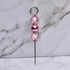 May include: A pink and silver beaded accessory with a metal pin. The accessory features three pink beads, a silver chain, and a small charm. The top has a silver ring.