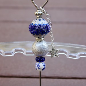 May include: A silver and blue beaded bottle stopper with a star charm. The stopper is on a clear acrylic stand.