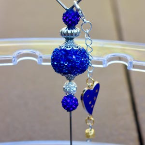 May include: A blue and silver charm with a heart-shaped top, a large blue rhinestone ball, a smaller blue rhinestone ball, and a silver heart charm with blue stars. The charm is attached to a silver pin.