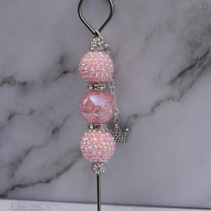 May include: A pink beaded accessory with a silver loop and a pointed end. The accessory features three pink, sparkly beads and a small silver crown charm. The beads are adorned with iridescent rhinestones.