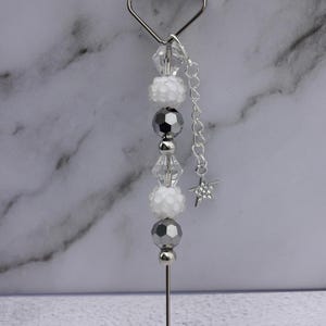 May include: Silver-toned metal heart-shaped topper with a beaded design. The beads are white, clear, and gray, with a small star charm hanging from a chain. The topper is designed to be inserted into a cake or other dessert.