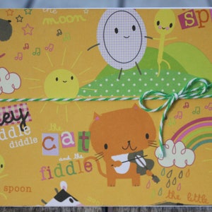 May include: A whimsical greeting card with a yellow background featuring cartoon illustrations of a cat playing a fiddle, a sun, and a rainbow. Text includes "Hey Diddle Diddle" and "The Cat and the Fiddle". Tied with green and white twine.