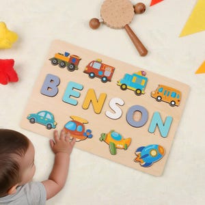 May include: A wooden name puzzle spelling "BENSON" in colourful letters, with vehicle shapes like a train, fire engine, car, helicopter, aeroplane, bus, and rocket. A wooden rattle is in the background. The puzzle is designed for young children.