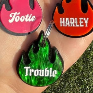 May include: A collection of five flame-shaped pet tags in various colors, including pink, orange, green, purple, and white. Each tag features a different pet name in a contrasting color, such as "Tootie" and "Harley". The tags are attached to silver key rings.