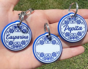 May include: Three round keychains with blue and white floral designs. Two keychains have names, "Casparina" and "Pepita", in blue script. The third keychain has a blank space for personalization. Each keychain has a silver keyring.
