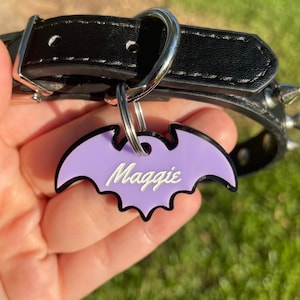 May include: A black leather dog collar with silver hardware and metal spikes. A purple bat-shaped tag with the name "Maggie" in white script hangs from the collar. The collar is shown against a blurred green background.