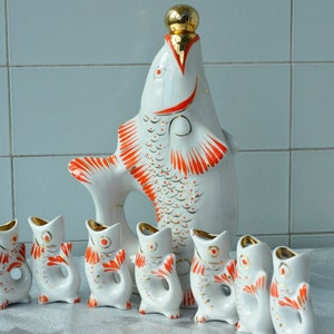 Peut inclure: Un ensemble de verres à liqueur en porcelaine blanche en forme de poisson et une bouteille plus grande en forme de poisson. Les poissons sont décorés d'accents rouges et dorés.