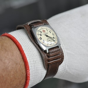 Puede incluir: Un reloj de pulsera plateado con una esfera blanca y una correa de cuero negra. La esfera del reloj tiene marcadores de hora en números romanos negros y una estrella roja en la posición de las 12 en punto. La esfera del reloj también tiene el texto "СССР" en escritura cirílica.