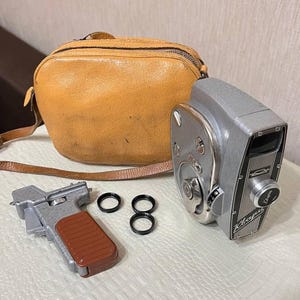 May include: A vintage silver camera with a brown leather case and strap. The camera has a black and silver design with a lens and dials. Also included is a silver grip with a brown handle and two black rings.
