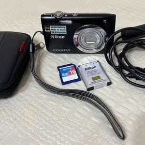 May include: A black Nikon Coolpix digital camera with a wrist strap, a black carrying case, a memory card, a battery, and a USB cable. The camera has a silver lens and the text "COOLPIX" on the front.