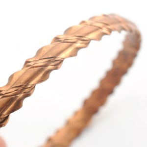 May include: A copper bangle bracelet with a braided design. The bracelet is smooth and shiny.