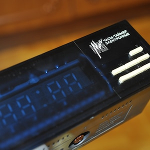 May include: A black digital clock with a blue display showing the time 12:21. The clock has a white label that reads "Часы-таймер Электронные".