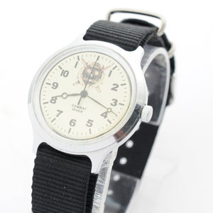 Puede incluir: Un reloj de pulsera plateado con una correa de nylon negra. La esfera del reloj es blanca con números y manecillas negros. La esfera del reloj tiene un gráfico de un buzo con espadas cruzadas y las palabras "Combat Diver".