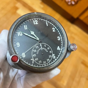 May include: A vintage, round, black-faced watch with white numerals and hands. The watch has a smaller subdial and a red button on the side. The watch is held by a gloved hand.