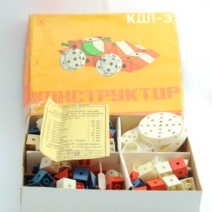 May include: A vintage plastic toy construction set in an orange box with the text "КДП-3 КОНСТРУКТОР" on the front. The box contains a variety of colorful plastic pieces, including wheels, blocks, and other shapes. The box also includes a sheet of instructions with a list of parts and their quantities.