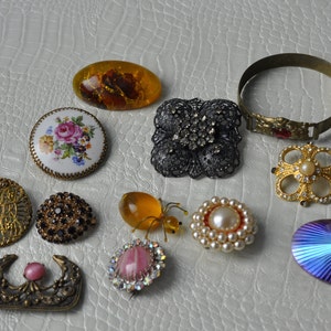 May include: Assortment of vintage brooches and jewelry pieces, including a floral porcelain brooch, a gold-tone filigree brooch, a black and silver brooch with rhinestones, a gold-tone brooch with pearls, a gold-tone bracelet, a blue iridescent brooch, a pink and clear crystal brooch, a yellow amber brooch, and a brown and gold brooch.