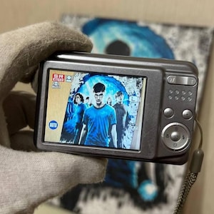May include: A silver digital camera showing a film scene on its screen. The screen displays a group of people, with a man in a blue shirt in the centre. The camera has several buttons and a strap.