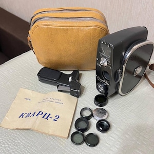 Może przedstawiać: Zabytkowa czarna kamera filmowa Quartz-2 ze skórzanym etui, wizjerem i kilkoma obiektywami. Aparat ma srebrne wykończenia i brązowy skórzany pasek. Dołączona jest również instrukcja z tekstem cyrylicą.