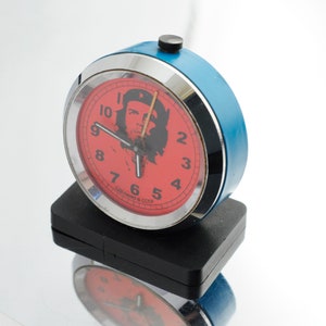 May include: A vintage blue alarm clock with a red face featuring a black silhouette of Che Guevara. The clock has a silver bezel and black base. The clock face has black numerals and hands.